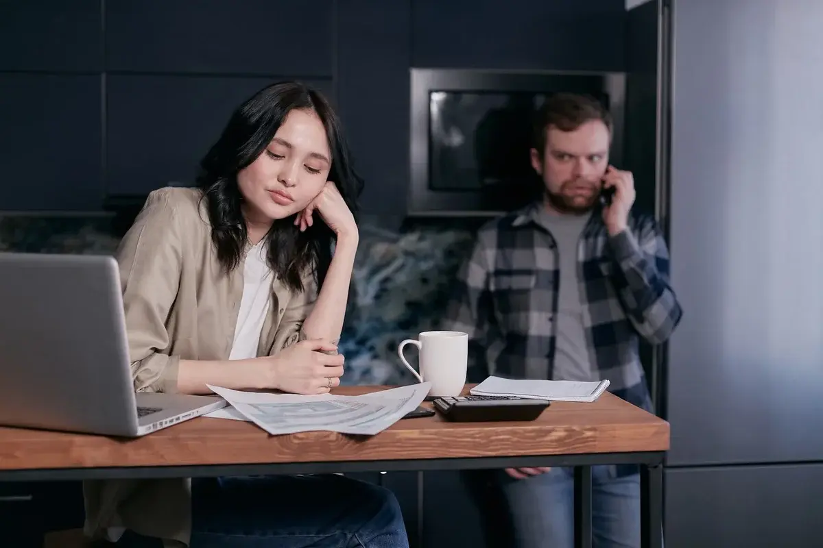 A couple reviewing financial papers at a kitchen table, with the man on a phone in the background.