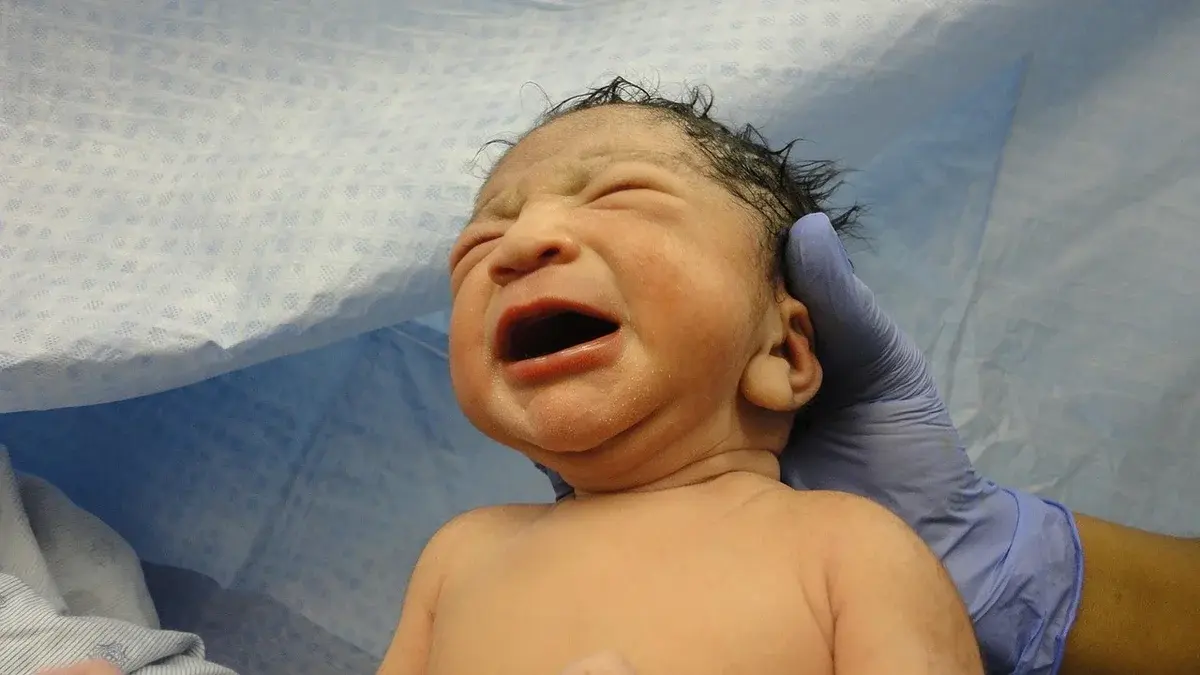 A crying newborn baby being held by a gloved hand in a hospital setting.